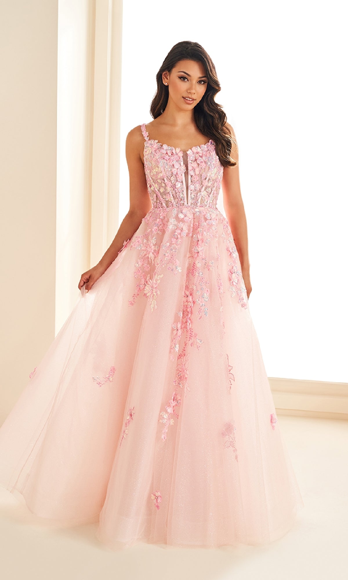 A woman stands in a well-lit room wearing the Ellie Wilde EW36213, a sleeveless, floor-length pink prom dress featuring floral lace appliques and a corset bodice.